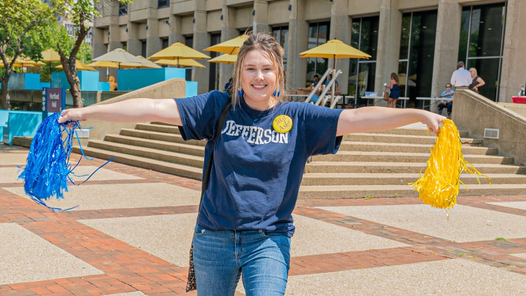 female student wearing a Jefferson shirt with pom poms, standing at a Jefferson building