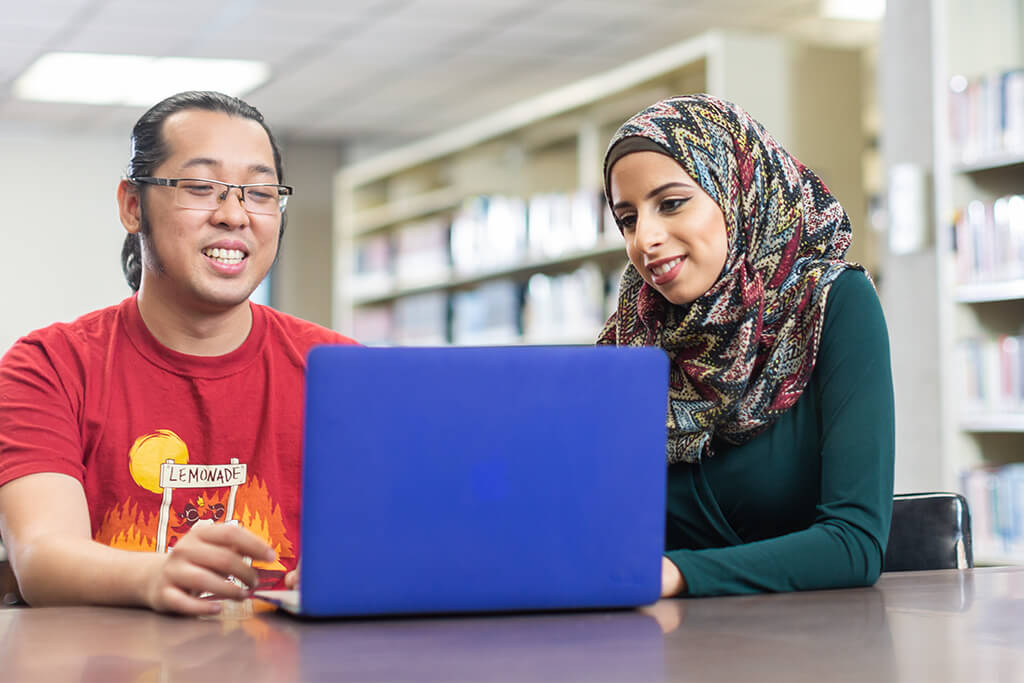 two students sitting at table and looking at laptop